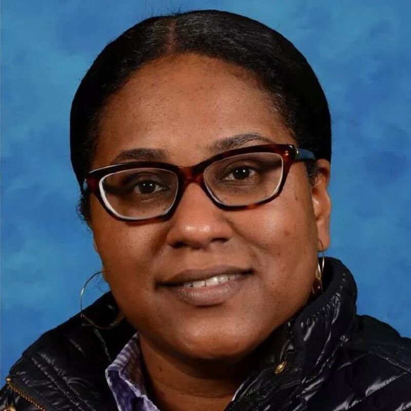 The image shows a woman with dark skin wearing glasses and a black jacket. She has dark hair pulled back and is looking at the camera. The background is a solid blue color. She appears to be in a professional setting.